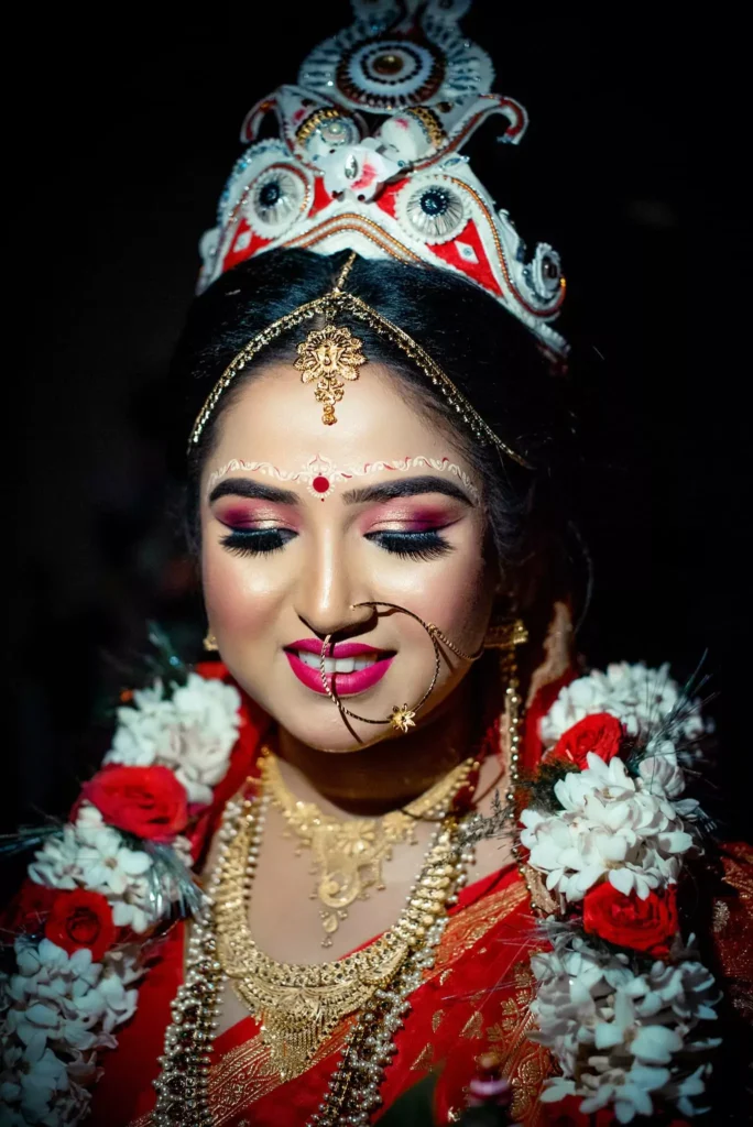 The bride, adorned in traditional Bengali bridal attire with a prominent mukut (headgear) and layers of gold jewelry, smiles gently with her eyes closed, showcasing her bright pink lipstick and beautifully crafted eye makeup.