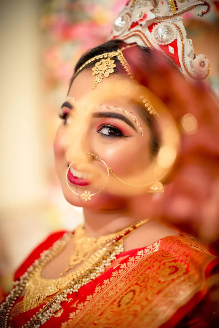 A close-up of a bride in traditional Bengali wedding attire, showcasing intricate face art and jewelry. Her eyes are highlighted with bold eye makeup, and she wears a bright smile, partially obscured by a blurred object in the foreground.