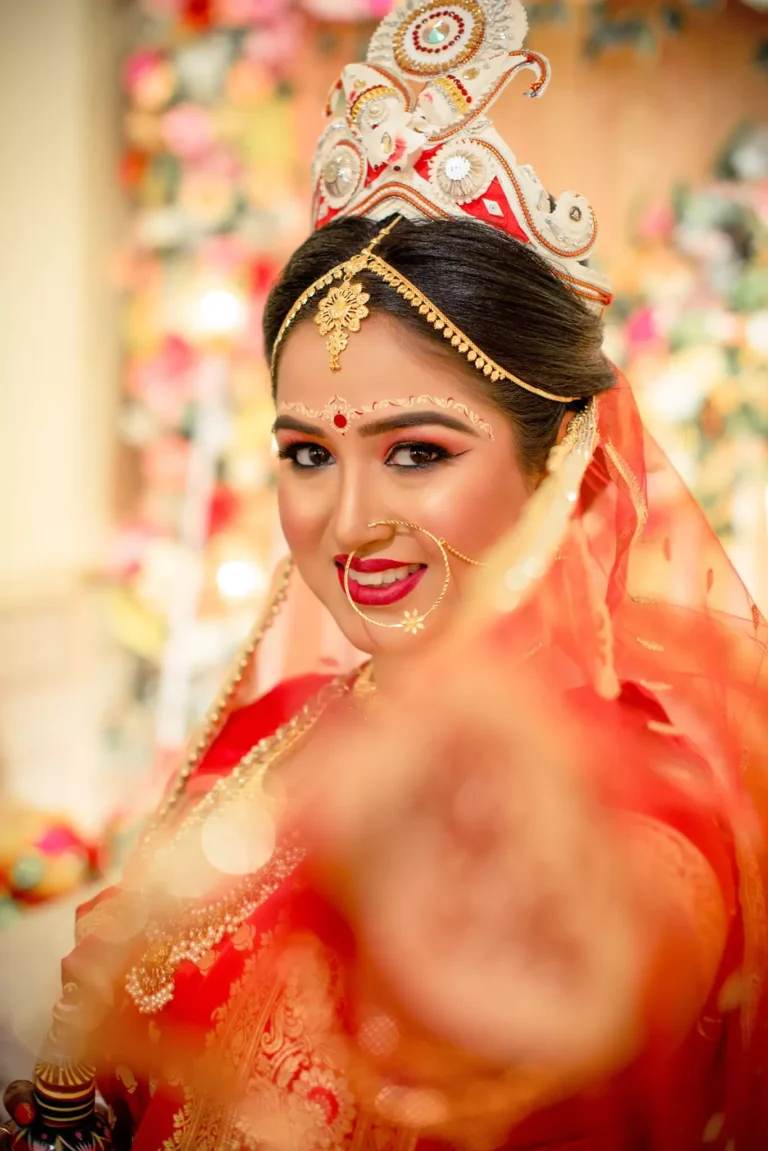 The bride, dressed in a vibrant red saree and adorned with traditional jewelry, poses with a confident smile. Her outstretched arm and blurred foreground create a dynamic and engaging composition.