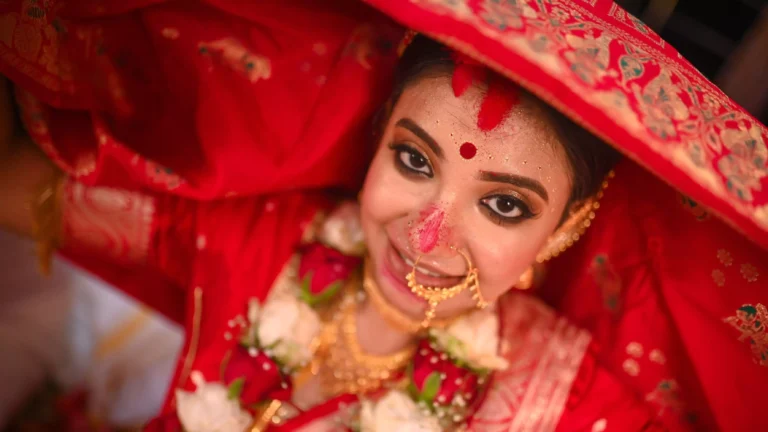 Close-up of a bride in traditional Indian wedding attire, smiling under a red veil. She wears intricate gold jewelry, including a nose ring and necklace, with red and white floral garlands. Her makeup features bold eye makeup and red sindoor on her forehead, symbolizing her marital status.