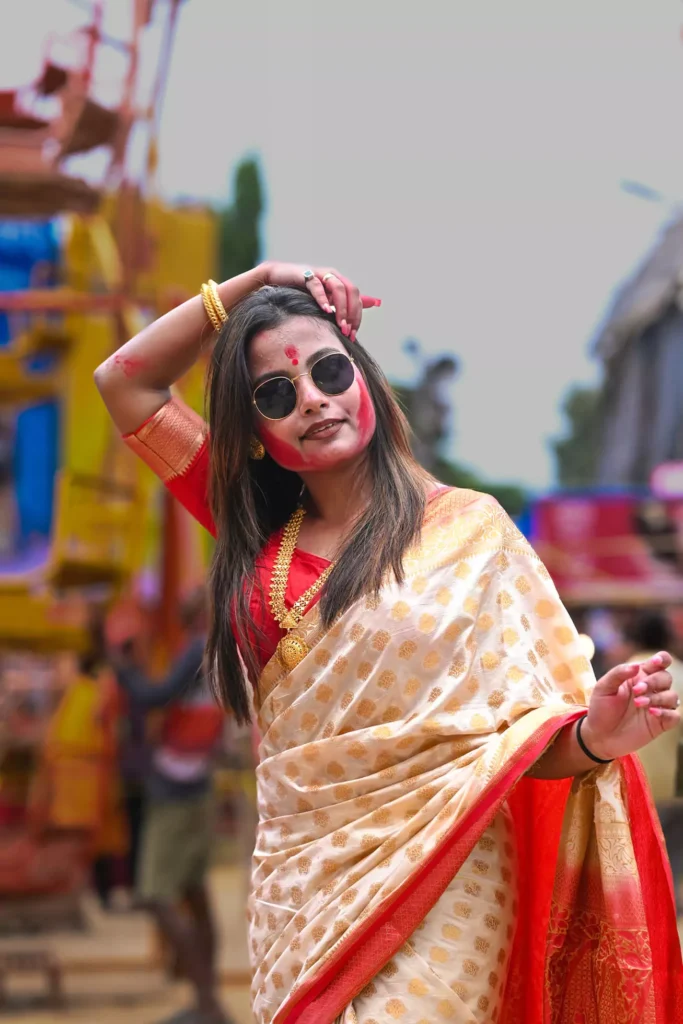 Woman in traditional saree with red and white face paint, wearing sunglasses and posing during a festival.