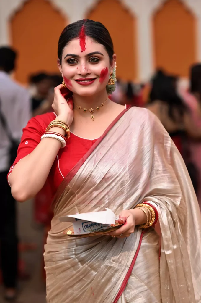 Woman in a cream saree with red and gold accents, holding a tray during a religious celebration.