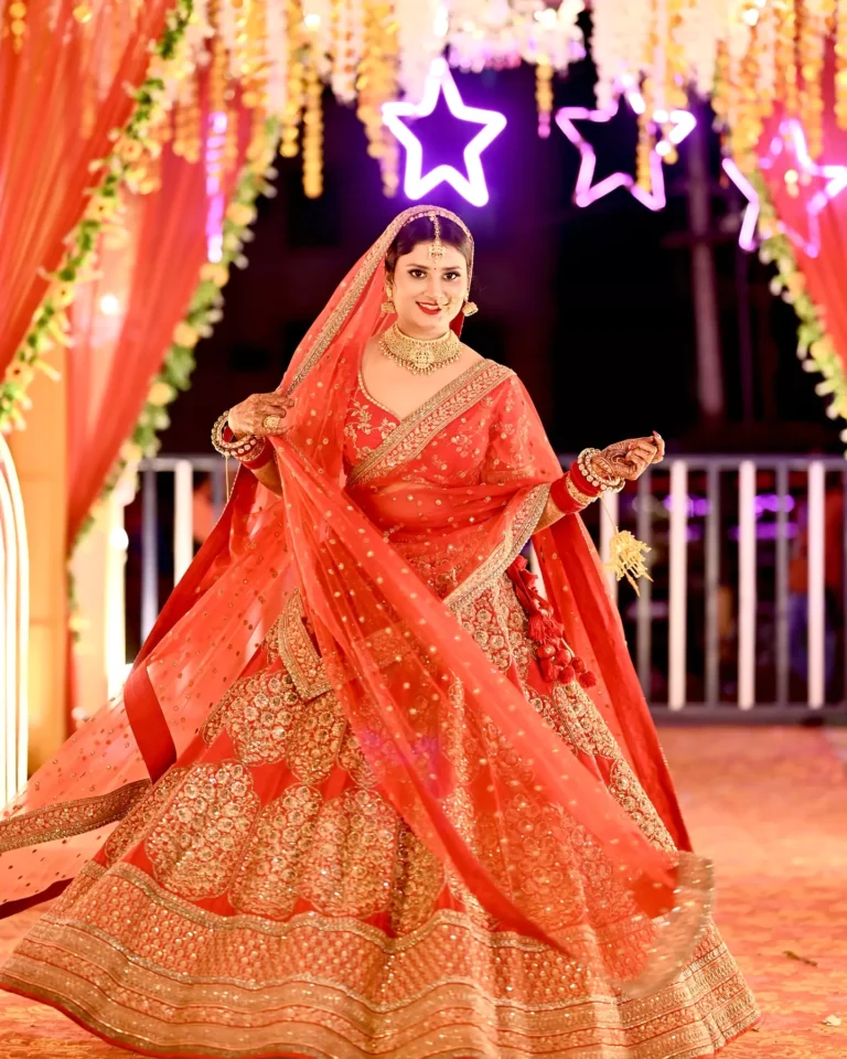 Bride wearing a red lehenga, smiling while posing under decorative lights and floral arrangements.