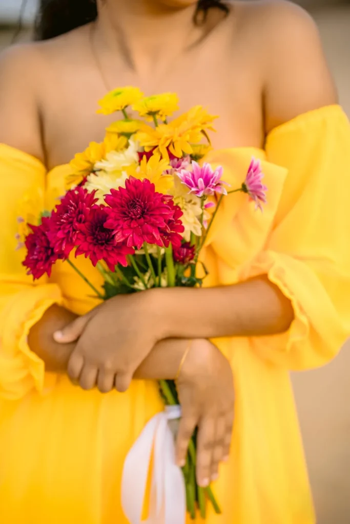 A beautiful young woman in a yellow dress holds a colourful bouquet of flowers, showcasing her beauty.