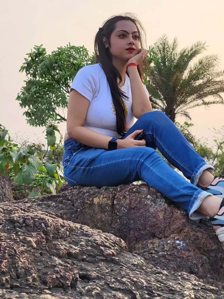Image is a portrait-style photograph featuring a woman sitting on a large rock outdoors. She has long, dark hair tied back, and her skin is light. She is wearing a white t-shirt and blue jeans, with white and black sneakers. Her left hand rests on her chin, and her right hand holds a black object, possibly a phone. She is wearing a black wristwatch on her left wrist and a red bracelet on her right wrist. In the background, there are green trees and a palm tree, with a clear sky above. The overall composition is natural and relaxed, capturing a moment of contemplation.