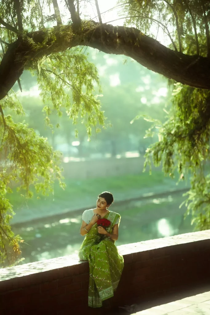 Woman in green saree sitting under a tree near a lake.