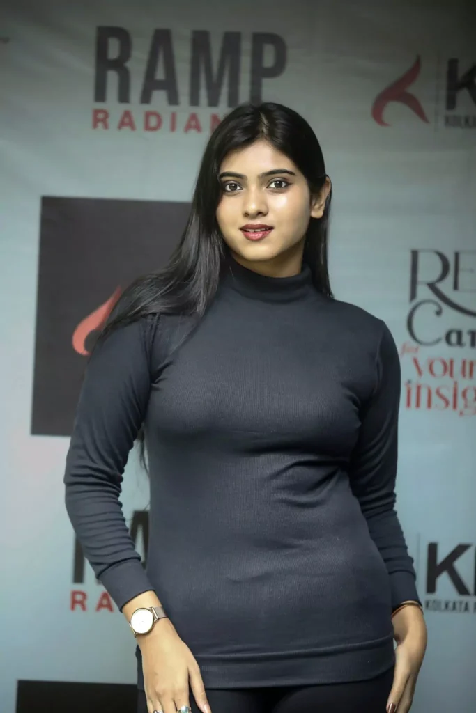 Elegant female model standing straight in black fitted dress with soft smile, in front of fashion event backdrop.