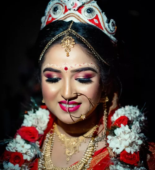 The bride, adorned in traditional Bengali bridal attire with a prominent mukut (headgear) and layers of gold jewelry, smiles gently with her eyes closed, showcasing her bright pink lipstick and beautifully crafted eye makeup.