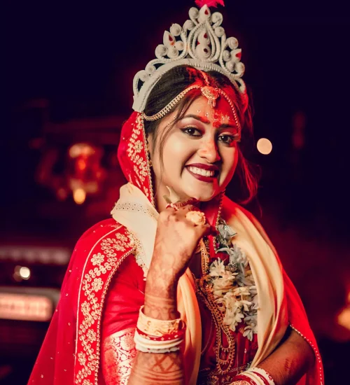 Bengali bride smiling with hands covered in mehndi, wearing red bridal saree.